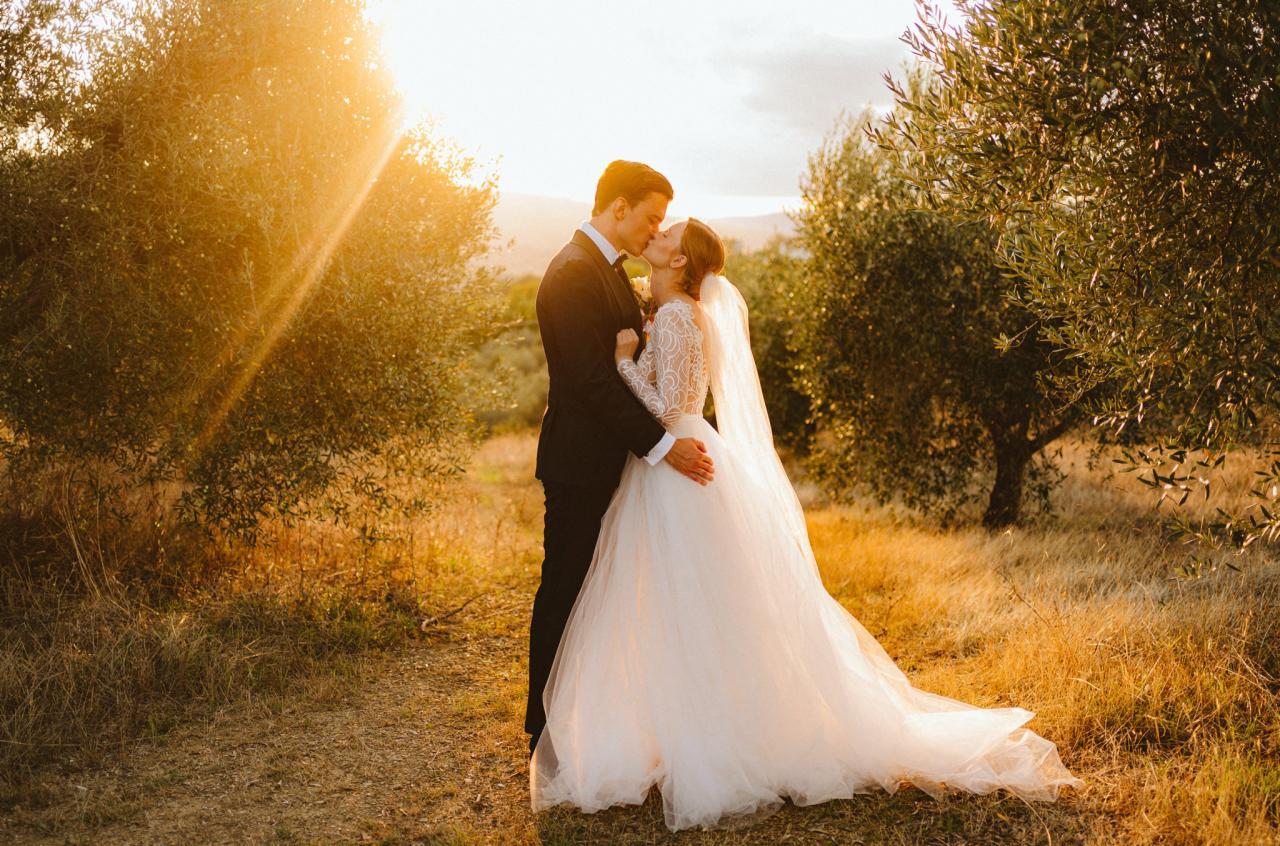 Para młoda w objęciach całuje się na tle drzew oliwnych w złocistym świetle zachodzącego słońca, ona w koronkowej sukni z welonem, on w czarnym garniturze.