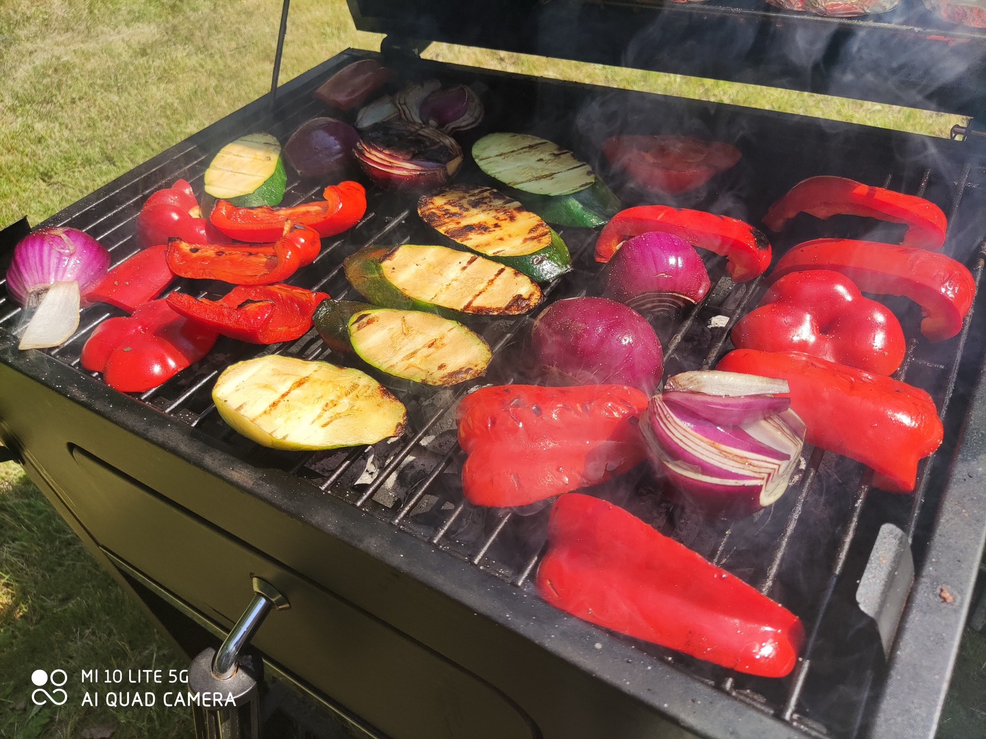 Grillowane warzywa: czerwona papryka, cebula i cukinia na ruszcie, z widocznymi śladami grillowania i delikatnym dymem.