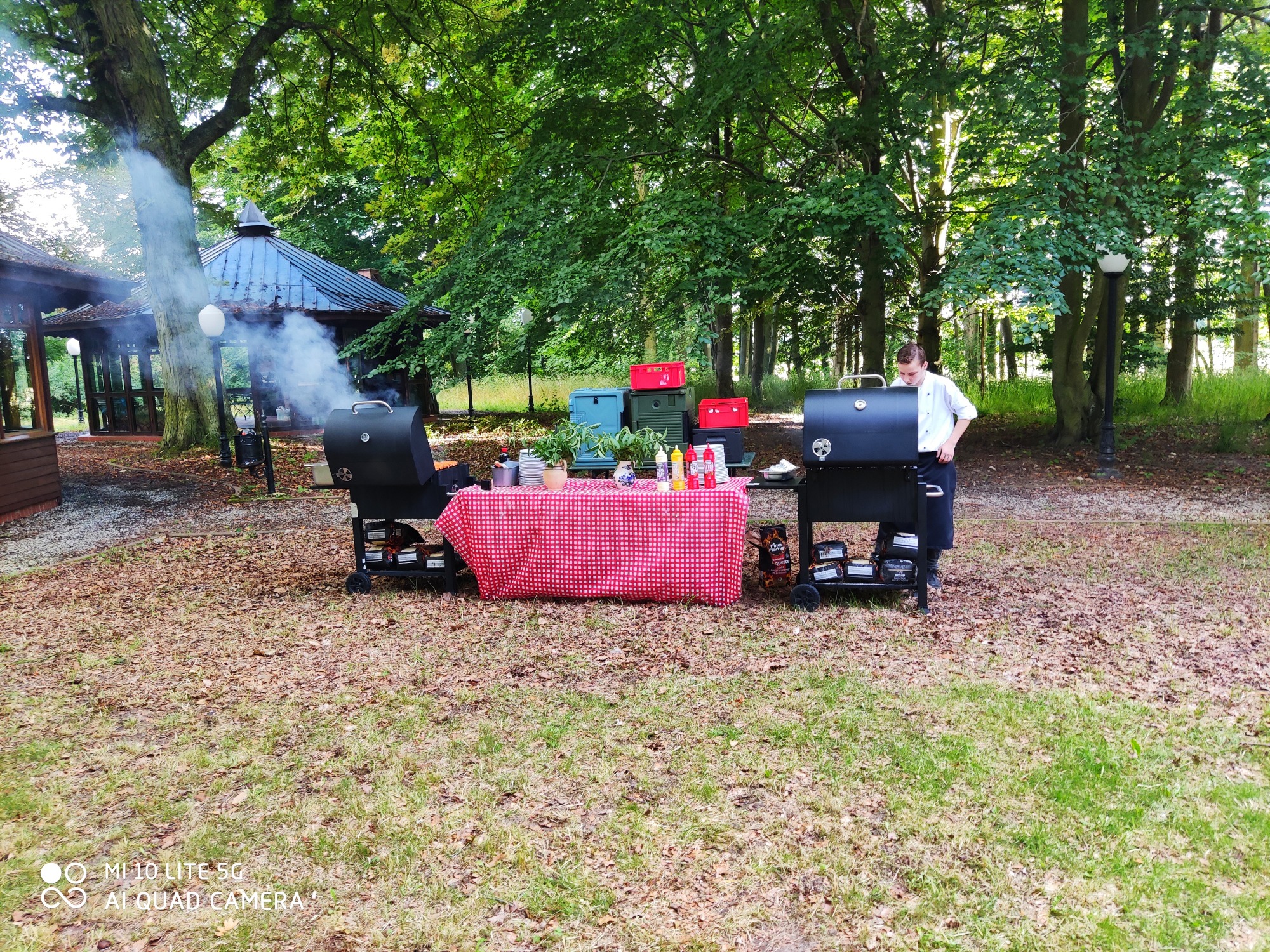 Przygotowania do grilla na zewnątrz: kucharz w białej koszuli przy czarnym grillu, stół z czerwono-białym obrusem w kratę, sosy, naczynia, dekoracje roślinne, w tle altana i dym z grilla.