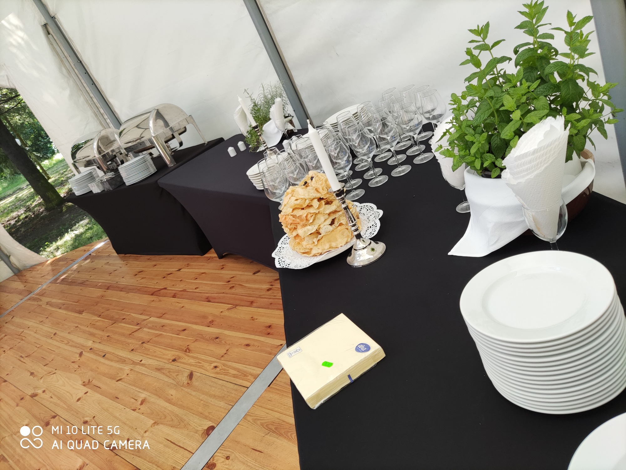 Przygotowany stół bufetowy z czarnym obrusem w namiocie: podgrzewacze do potraw, stosy białych talerzy, piramida z ciasta na srebrnym stojaku, świece, szklanki i doniczka z miętą.