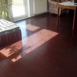 Ciemna, lakierowana podłoga z widocznymi promieniami słońca wpadającymi przez okno, częściowo zasłonięte firanką z wzorem, w tle fotel, stół i roślina doniczkowa.