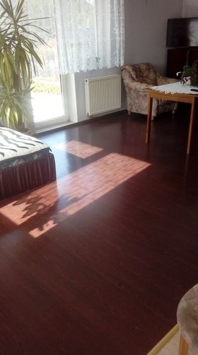 Ciemna, lakierowana podłoga z widocznymi promieniami słońca wpadającymi przez okno, częściowo zasłonięte firanką z wzorem, w tle fotel, stół i roślina doniczkowa.