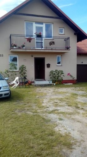 Dwupiętrowy dom z balkonem z czarną balustradą, na którym stoją doniczki z czerwonymi kwiatami, widok z zewnątrz, trawnik z przodu.