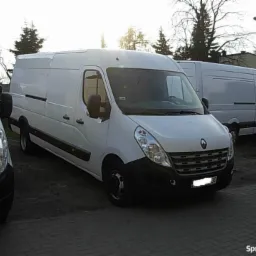 Białe busy Renault Master zaparkowane na brukowanym placu, widoczne z przodu, jeden z lekko otwartymi drzwiami kierowcy.