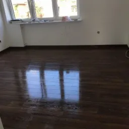 Pokój po remoncie z ciemną, błyszczącą podłogą z paneli, odbijającą światło z okna. Ściany białe, częściowo widoczne kable przy podłodze.