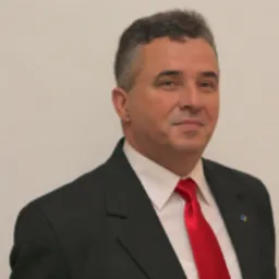 Portret mężczyzny w garniturze z czerwoną koszulą i krawatem, uśmiechającego się do kamery na jasnym tle.