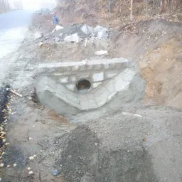 Konstrukcja przepustu drogowego w trakcie budowy, z widoczną rurą i elementami kamiennymi, częściowo obłożona betonem, w tle droga i drzewa.