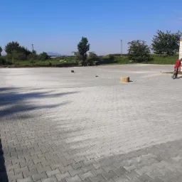 Pracownik zagęszcza wibratorem świeżo ułożoną, szarą kostkę brukową na placu, widoczna krawędź z obrzeży, w tle budynek w stanie surowym i zieleń.