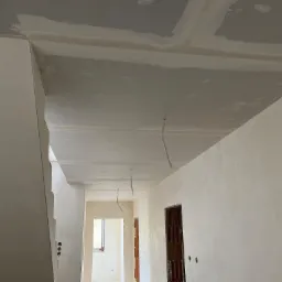 Wnętrze w trakcie remontu: sufit z płyt karton-gips, świeżo szpachlowane ściany z widocznymi przewodami elektrycznymi, oraz fragmenty muru z cegły w otworach drzwiowych.