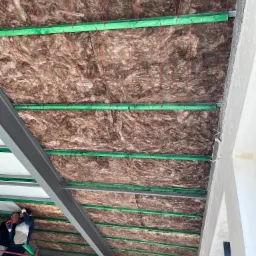 Montaż izolacji z wełny mineralnej na suficie, widoczne zielone listwy mocujące i czerwone sznurki, dwóch pracowników na drabinie w lewym dolnym rogu.