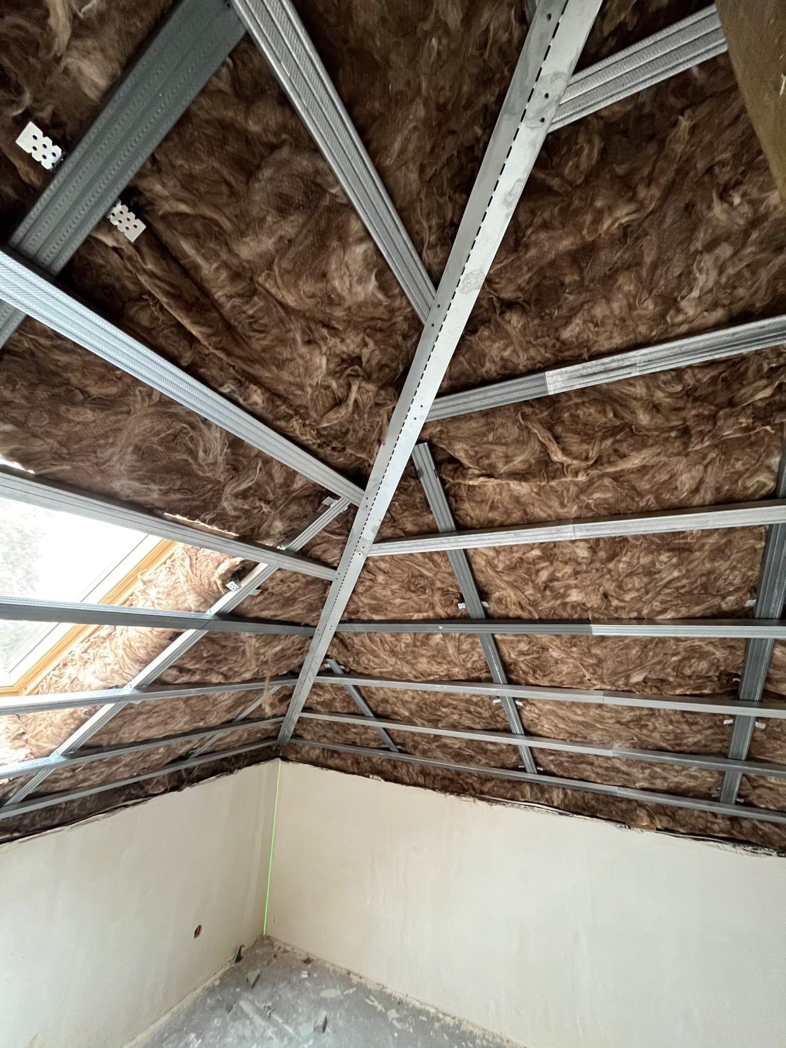 Montaż metalowej konstrukcji sufitu podwieszanego z wełną mineralną w przestrzeni poddasza, widoczna zielona linia lasera poziomującego na ścianie.