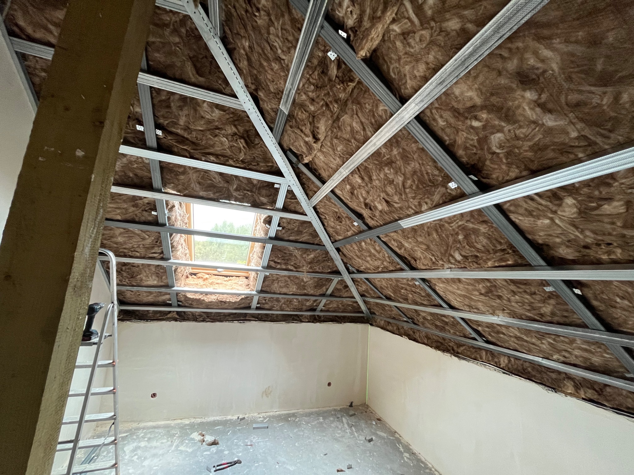 Poddasze w trakcie ocieplania: widoczna wełna mineralna między metalowymi profilami konstrukcyjnymi dachu, drabina oparta o ścianę, surowe ściany i podłoga w pomieszczeniu.