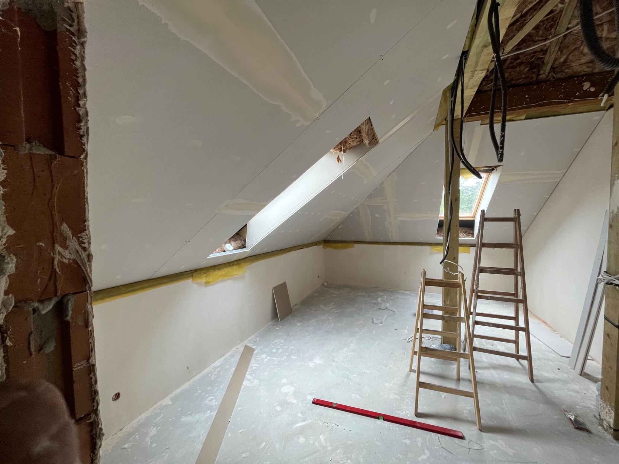 Poddasze w trakcie prac wykończeniowych: zamontowane płyty na skosach, widoczne ocieplenie, dwie drewniane drabiny, poziomica na podłodze, surowa ściana z cegły.
