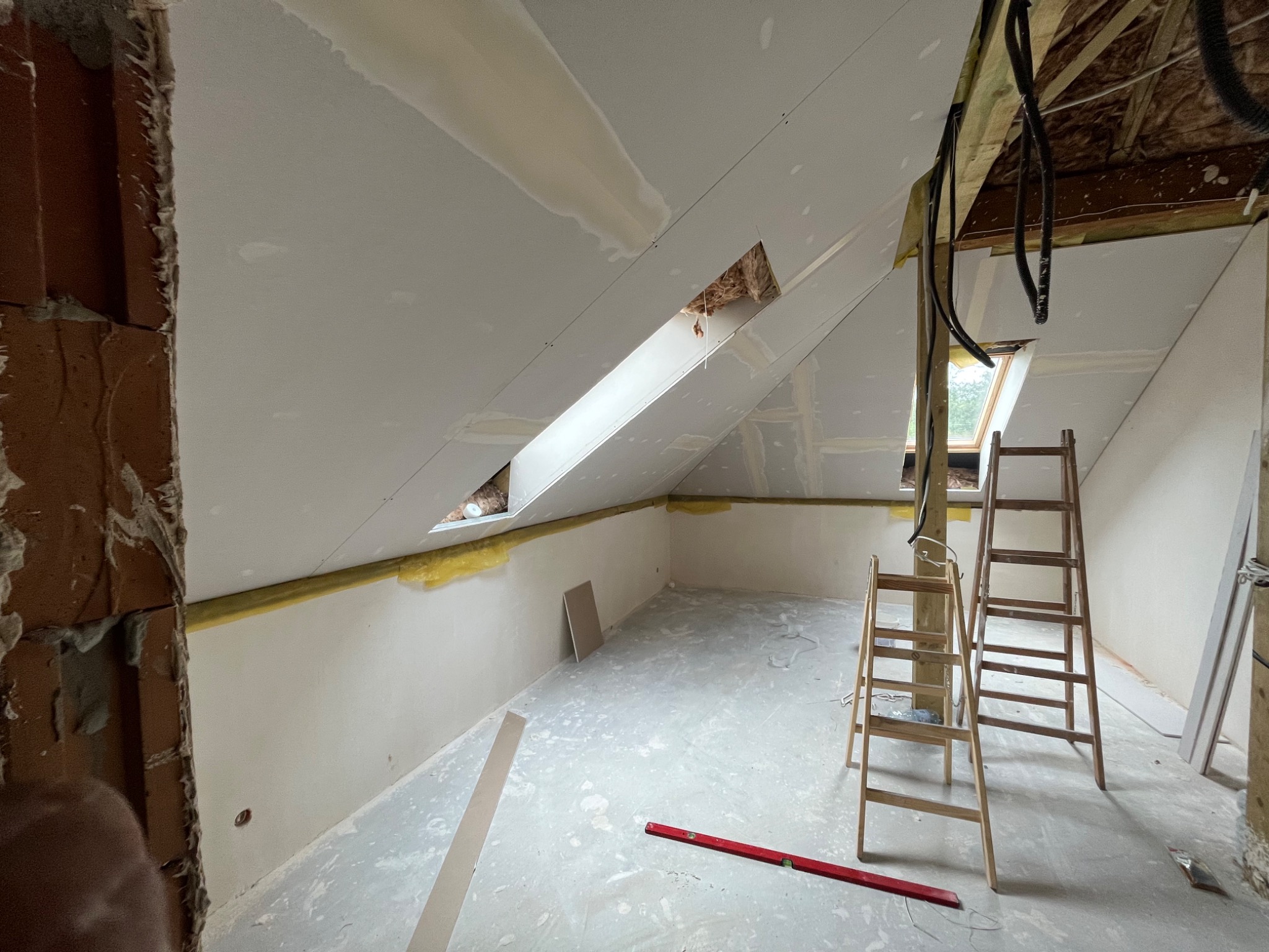 Poddasze w trakcie wykańczania: zamontowane płyty gipsowo-kartonowe na skośnym suficie i ścianach, widoczne otwory okienne, częściowo ocieplone ściany, dwie drewniane drabiny, czerwona poziomica...