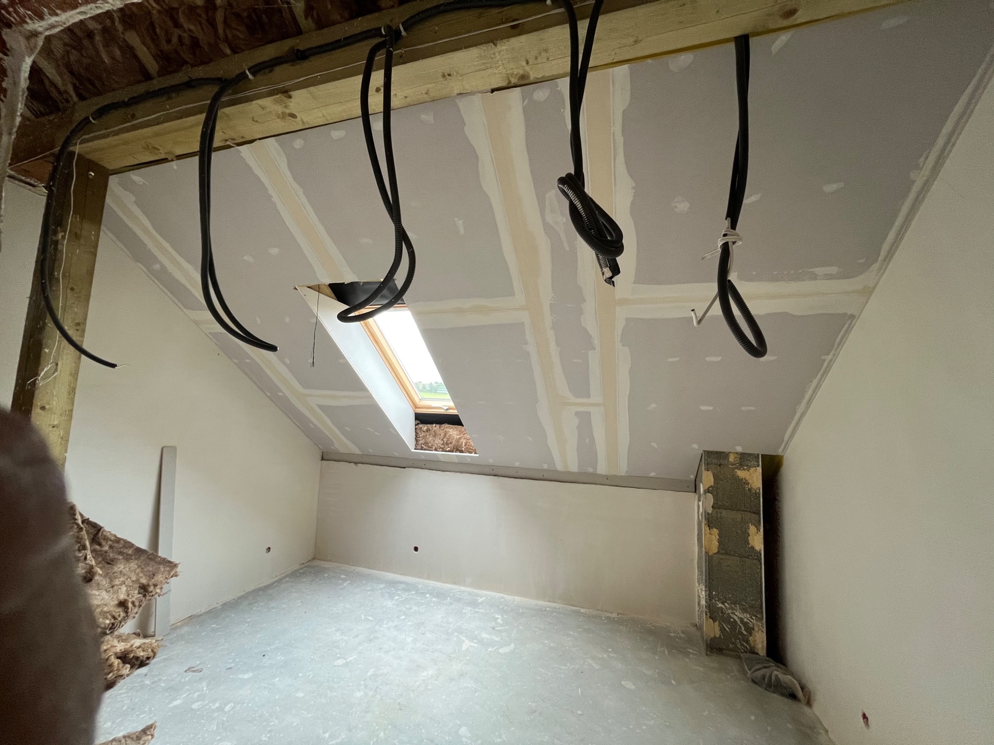 Poddasze w trakcie wykańczania: widoczna konstrukcja drewniana, szare płyty na suficie z widocznymi łączeniami, czarne kable zwisające z sufitu, okno dachowe, fragment ściany z cegły, surowa podłoga.