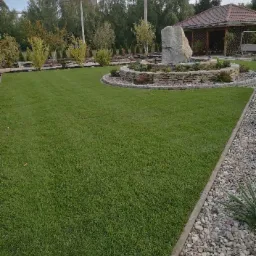 R&oacute;wny trawnik z centralnie umieszczoną kaskadą z kamienia i roślin na tle drzew i altany.