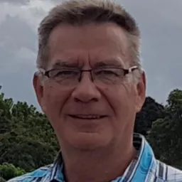 Portret uśmiechniętego mężczyzny w okularach i koszuli w paski, z drzewami i pochmurnym niebem w tle.