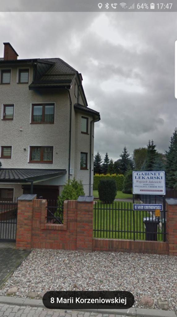 Budynek gabinetu lekarskiego Wojciecha Jakowickiego z tablicą informacyjną i adresem przy ulicy Marii Korzeniowskiej, widok z zewnątrz, pochmurne niebo.