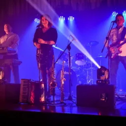 Zespół muzyczny na scenie podczas występu: wokalistka, gitarzysta, klawiszowiec, saksofon i akordeon w niebieskim oświetleniu.