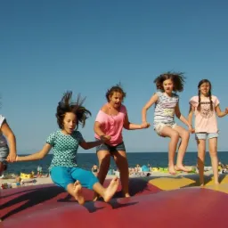 Grupa dziewcząt skacze trzymając się za ręce na dużej czerwono-żółtej dmuchanej poduszce na plaży, w tle morze i opalający się ludzie.