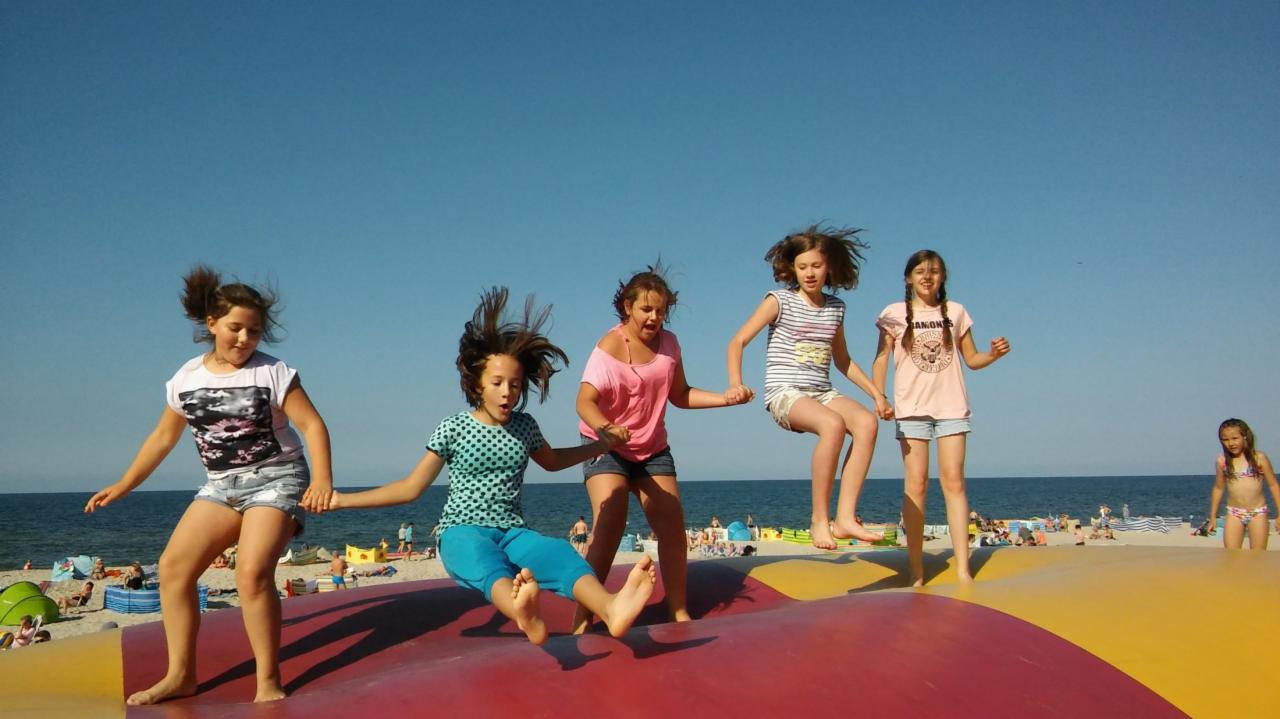 Grupa dziewcząt skacze trzymając się za ręce na dużej czerwono-żółtej dmuchanej poduszce na plaży, w tle morze i opalający się ludzie.