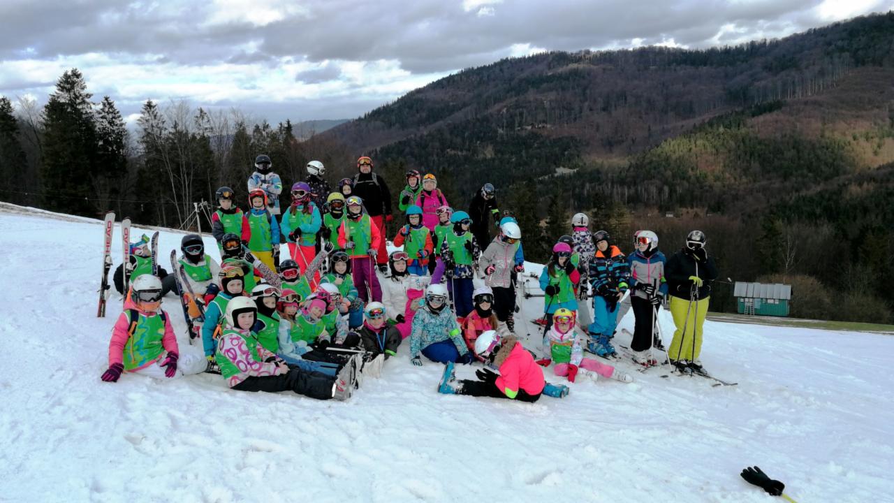 Grupa dzieci w kolorowych strojach narciarskich pozuje na śniegu, w tle zalesione wzgórze i pochmurne niebo.