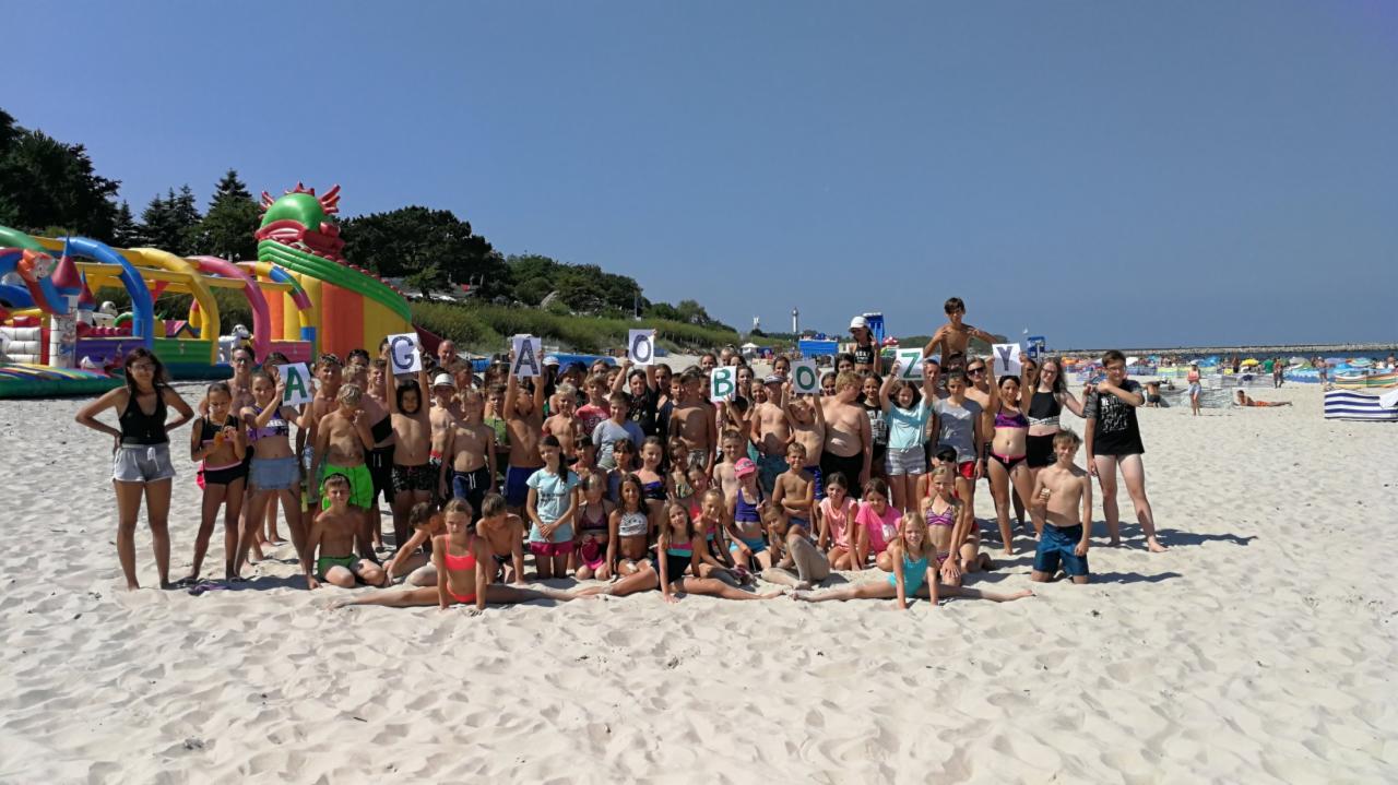 Duża grupa dzieci i młodzieży na piaszczystej plaży trzymająca kartki z literami układającymi się w napis 'GA OBOZY'. W tle widoczny dmuchany plac zabaw i morze.