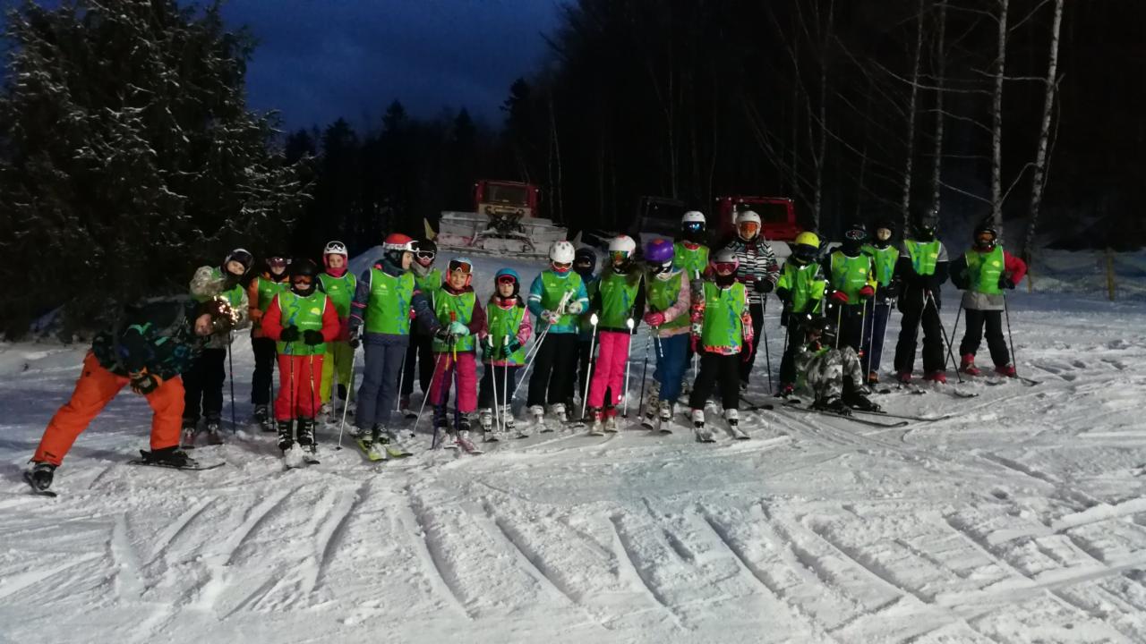 Grupa dzieci w zielonych kamizelkach na nartach, za nimi ratrak na ośnieżonym stoku o zmierzchu.