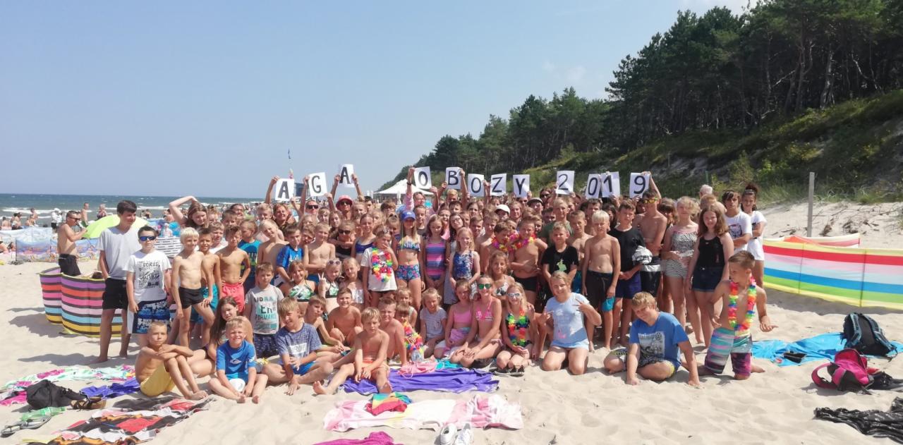 Duża grupa dzieci i młodzieży pozuje na plaży, trzymając litery układające się w napis 'AGA OBOZY 2019', w tle widać morze i las.