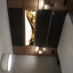 Nowoczesna sypialnia z podświetlanym panelem dekoracyjnym imitującym skałę nad łóżkiem, regulowana lampka nocna, biały materac w folii, minimalistyczna lampa wisząca.