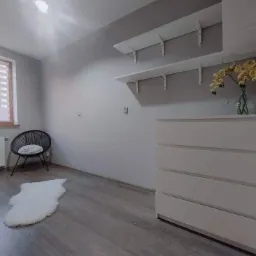 Jasnoszary pokój po malowaniu: białe ściany, drewniana podłoga, biała komoda z kwiatami, okno z żaluzjami, czarne krzesło z poduszką i biały dywanik.