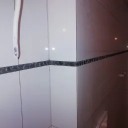 Wnętrze łazienki po remoncie z białymi płytkami i dekoracyjnym pasem z kamieniami, widoczny fragment instalacji hydraulicznej.