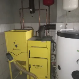 Żółty piec na pellet Pereko QmPell 35, zbiornik buforowy i instalacja CO w kotłowni z białymi kafelkami.