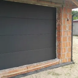 Ciemna, segmentowa brama garażowa w surowym stanie, widoczne cegły i niedokończone wykończenie otworu bramy.