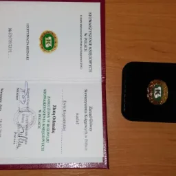 Dyplom Stowarzyszenia Księgowych w Polsce z Złotą Odznaką za zasługi, leżący obok otwartego pudełka z tą odznaką.