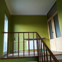Klatka schodowa z drewnianą balustradą i ścianami pomalowanymi na zielono, widoczny biały grzejnik i okno po lewej stronie.