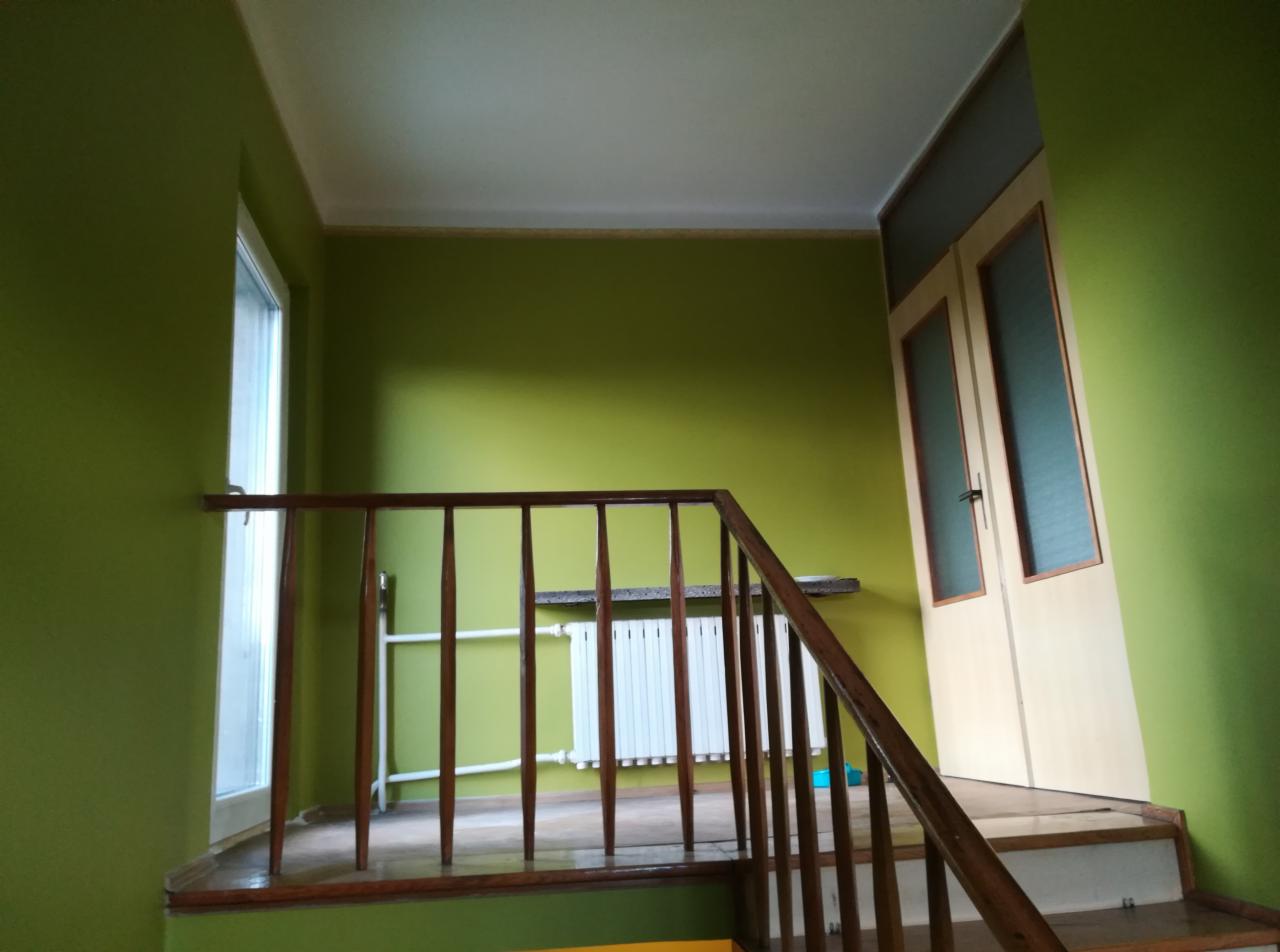 Klatka schodowa z drewnianą balustradą i ścianami pomalowanymi na zielono, widoczny biały grzejnik i okno po lewej stronie.