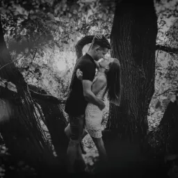 Zakochana para całuje się, otoczona drzewami w czarno-białej sesji plenerowej, z delikatnym światłem prześwitującym przez liście.
