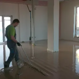 Pracownik w kombinezonie wyrównuje świeżo wylaną, brązową masę posadzkową przy użyciu listwy rozprowadzającej w jasnym pomieszczeniu z oknami.