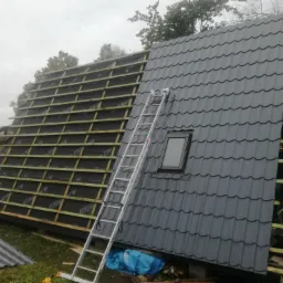 Budowa dachu: połowa konstrukcji z łatami i membraną, druga połowa pokryta szarą blachodachówką z oknem dachowym, drabina oparta o dach.