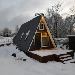 Nowoczesna altana w kształcie litery A z drewnianym tarasem, oświetlona ciepłym światłem, otoczona śnieżnym krajobrazem Grzmiącej podczas pochmurnego dnia.
