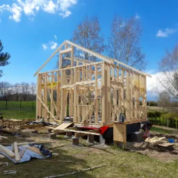 Konstrukcja szkieletowa domu jednorodzinnego w trakcie budowy, widoczne drewniane elementy nośne, narzędzia i materiały budowlane na trawie, błękitne niebo w tle.