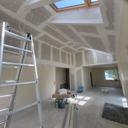 PRO-FACH - Wnętrze w trakcie prac wykończeniowych: widoczne płyty kartonowo-gipsowe na suficie i ścianach, aluminiowa drabina oparta o ścianę, okno dachowe wpuszczające światło, przygotowane materiały...