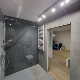 PRO-FACH - Nowoczesna kabina prysznicowa z deszczownicą i grzejnikiem drabinkowym na tle szarych płytek, widok na sąsiednie pomieszczenie z drewnianą podłogą i bałaganem w głębi.