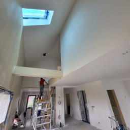 PRO-FACH - Wysoki pokój ze skośnym dachem w trakcie malowania. Malarz na rusztowaniu maluje ścianę pod oknem dachowym. Widoczne narzędzia malarskie i zabezpieczone elementy.