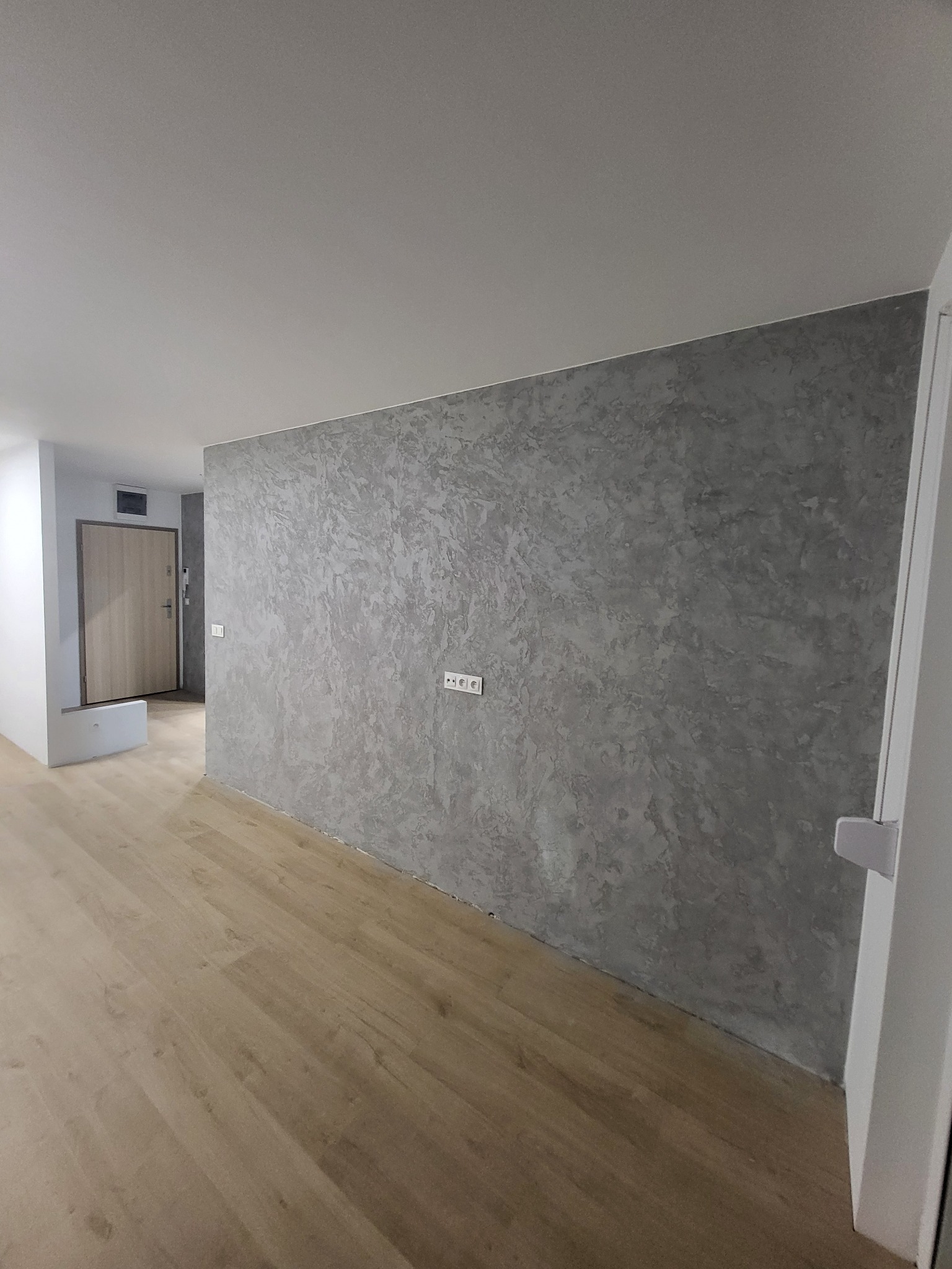 Pokój po remoncie, ściana pokryta dekoracyjną szarą fakturą przypominającą beton, jasna drewniana podłoga, widoczne gniazdka elektryczne.