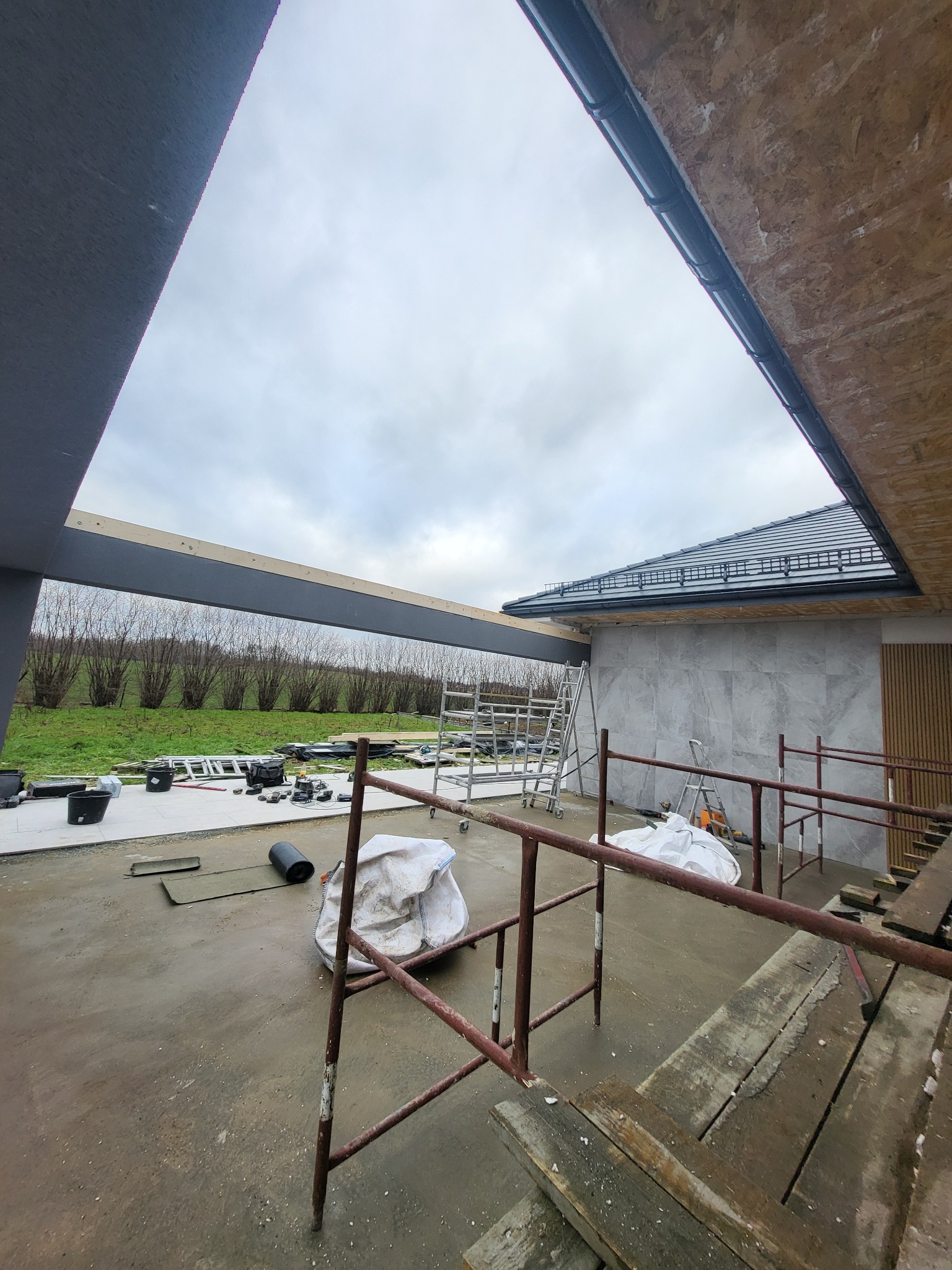 Budowa tarasu: widok na konstrukcję z rusztowaniem, materiałami budowlanymi i fragmentem domu w tle. Etap prac w toku, widoczne elementy wykończeniowe.