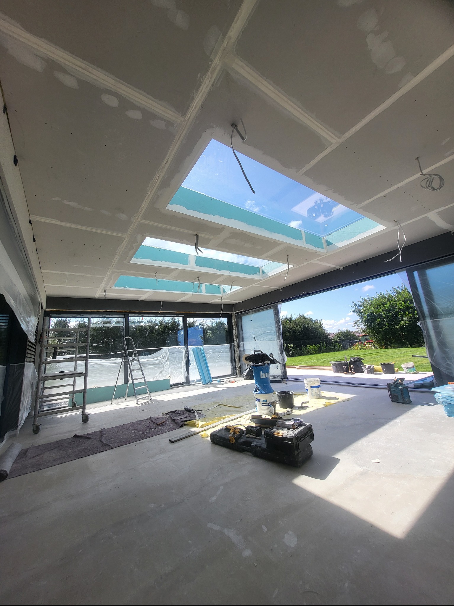 Wnętrze w trakcie montażu płyt gipsowo-kartonowych na suficie z oknami dachowymi. Widoczne narzędzia, drabina i materiały budowlane. Duże okna przesuwne z widokiem na zieleń.
