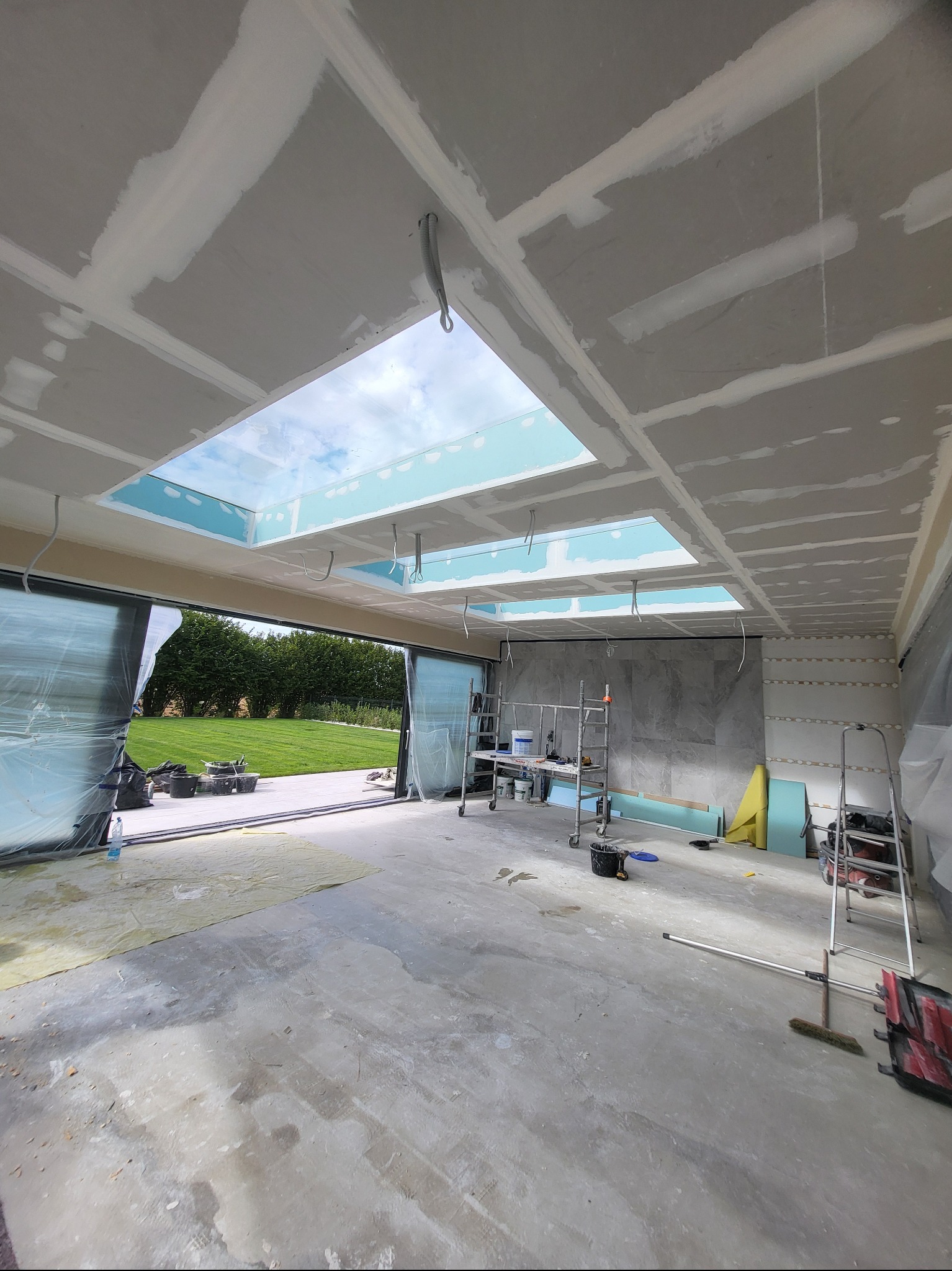 Wnętrze w trakcie montażu płyt karton-gips na suficie z oknami dachowymi. Widoczne rusztowania, narzędzia i otwarte przesuwne drzwi z widokiem na zewnątrz.