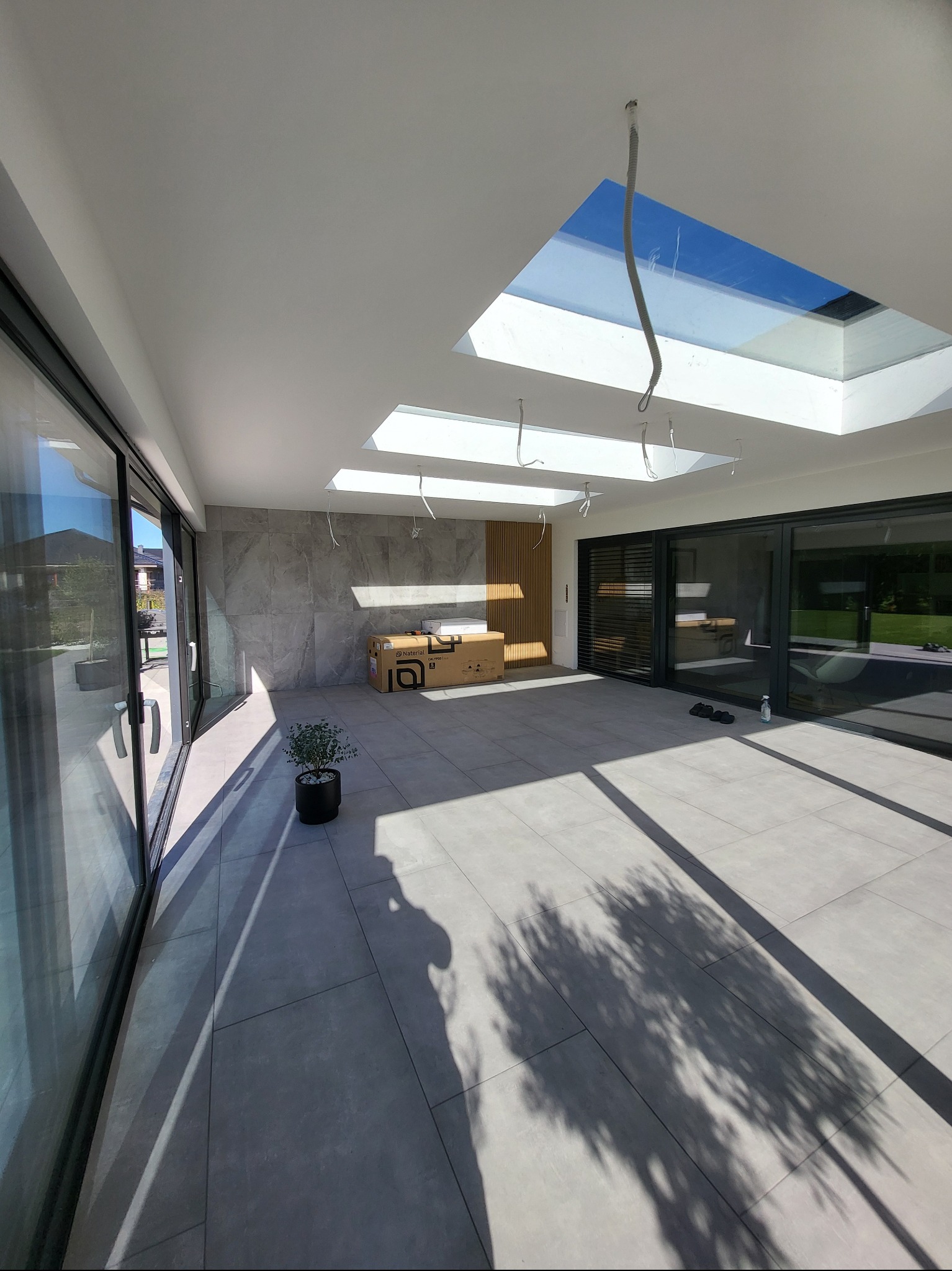 Jasne wnętrze z dużymi oknami i szarymi płytkami podłogowymi. Widoczne kartony, roślina w doniczce i fragmenty instalacji pod sufitem.